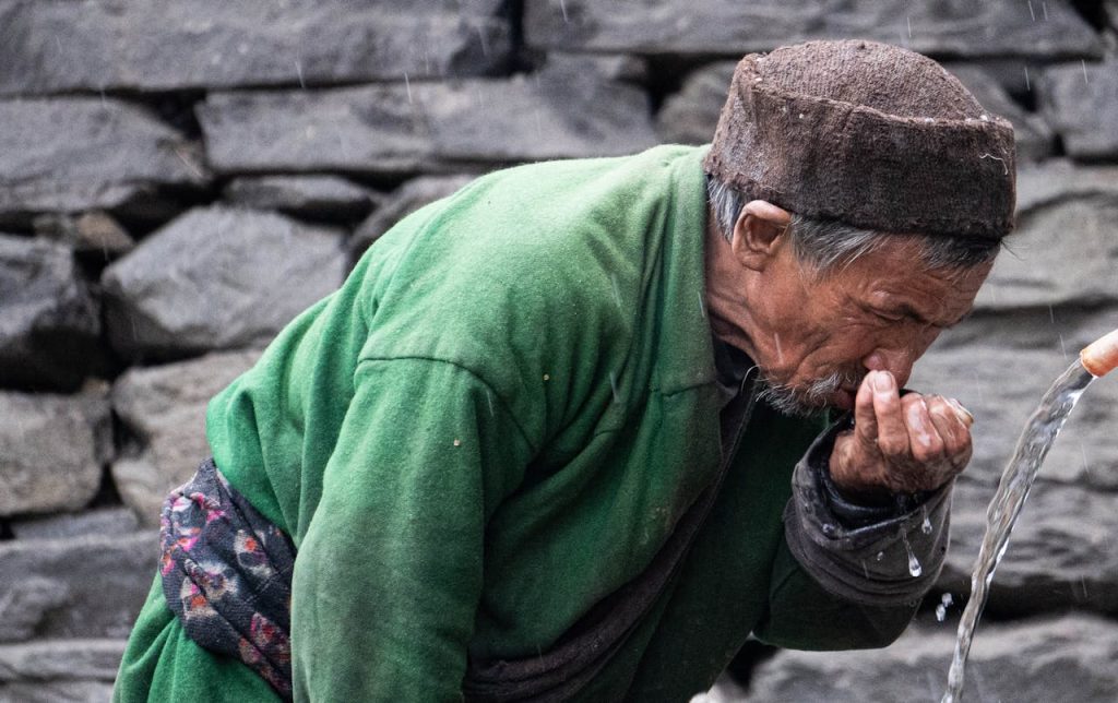 A senior man in traditional attire drinks from a stone fountain outdoors, showcasing cultural heritage.